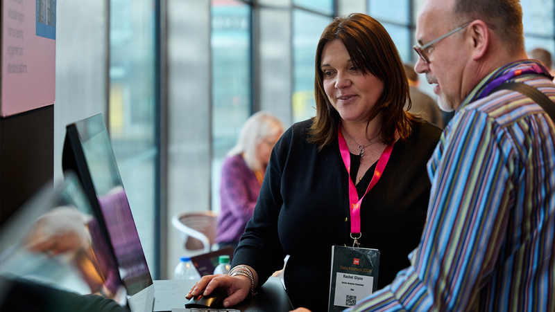 Two people at a counter looking at a computer during a professional event with large windows in the background.