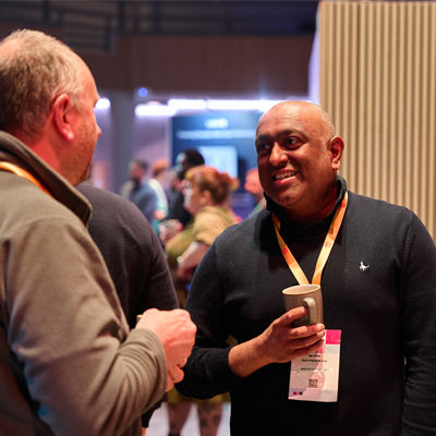 Two people talk at an event; one holds a cup and wears a lanyard. Others are in the background.