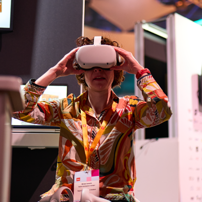 A person in a colorful shirt and orange lanyard uses a VR headset at an exhibition with screens and partitions in the background.
