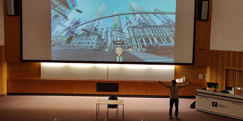 Professor Gary wears a VR headset while teaching a lecture. Behind him is a screen where he appears as a VR avatar.
