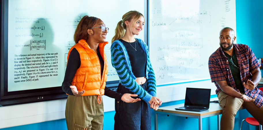 Technical college students standing at front of class presenting project with teacher looking on