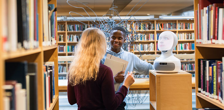 Students in the library talk next to a robot.