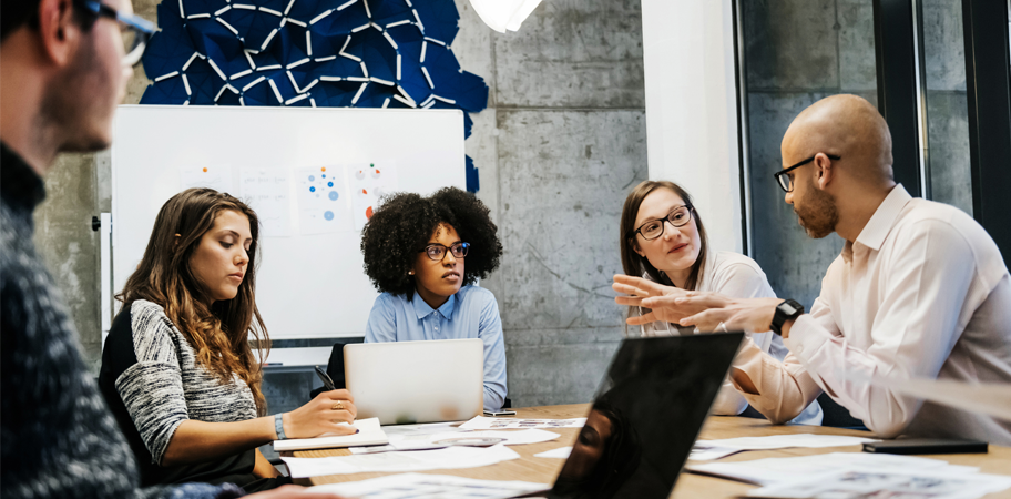 A group of colleagues sit around a table in a modern meeting room, discussing ideas with laptops, documents, and a whiteboard in the background.