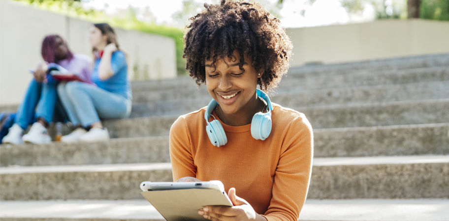 Latin young woman at school recess sitting on some stairs in the park with blue headphones around her neck and looking at a digital tablet while smiling.