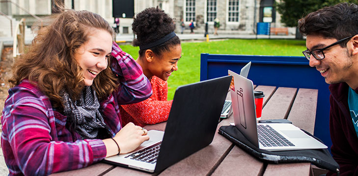 International students smile as they use their laptops on a bench outside on campus.