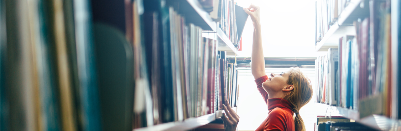 A woman selects a book from the top shelf of library book shelves.