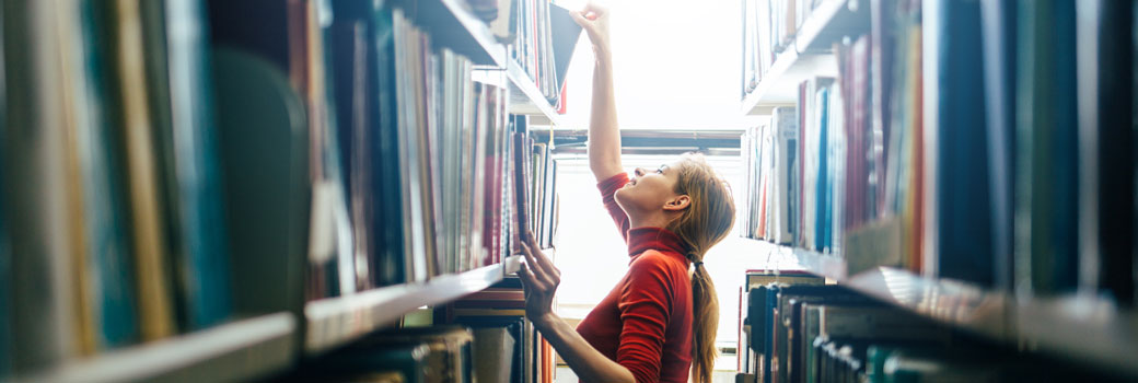 A woman selects a book from the top shelf of library book shelves.