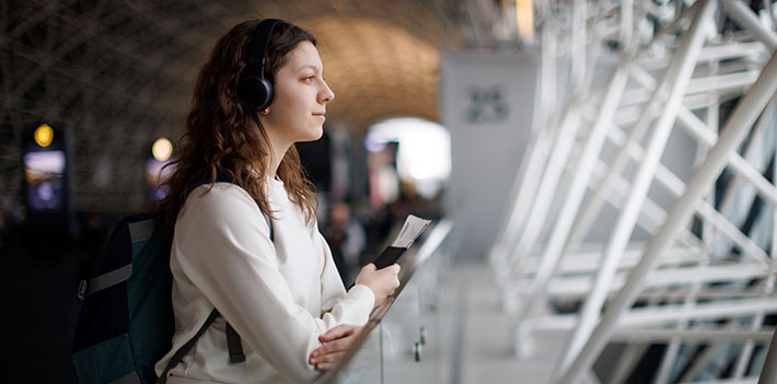 An international student arrives at a travel terminal. Holding her passport, ticket and backpack, she listens to the language of her new country in her headphones and looks apprehensively ahead to her future.