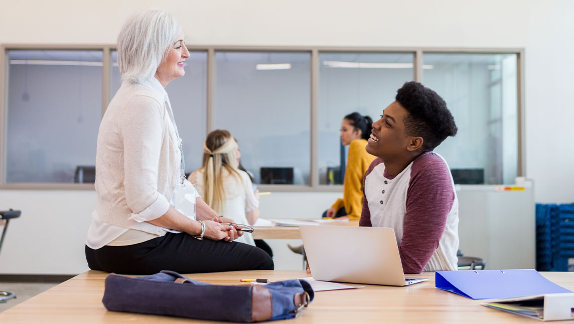 A student and teacher discuss work in a classroom.