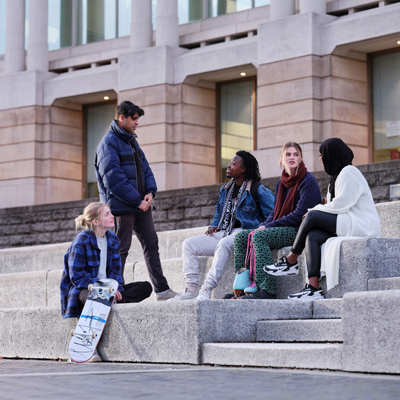 Four people sitting on stone steps outdoors, talking with another person standing nearby; a skateboard rests on the steps.