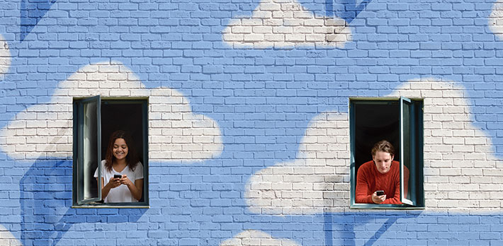 A building painted blue with clouds contains two open windows where students are using their mobile phones.