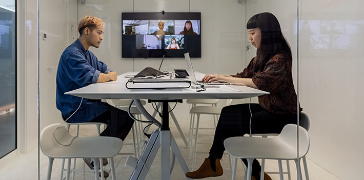 Two colleagues work in a hybrid meeting room with colleagues calling in remotely on a screen at the back.