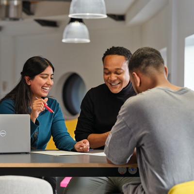 Three people sitting at a table in a modern office, discussing documents with a laptop nearby.