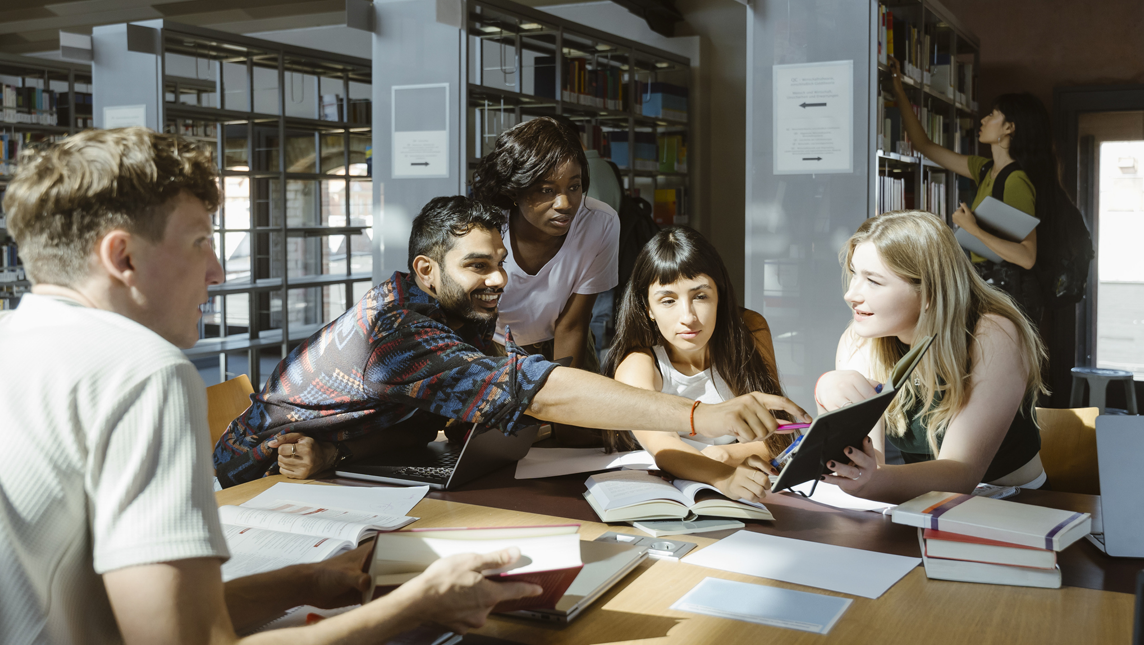Smiling man pointing at book while doing group study with male and female friends in library at university