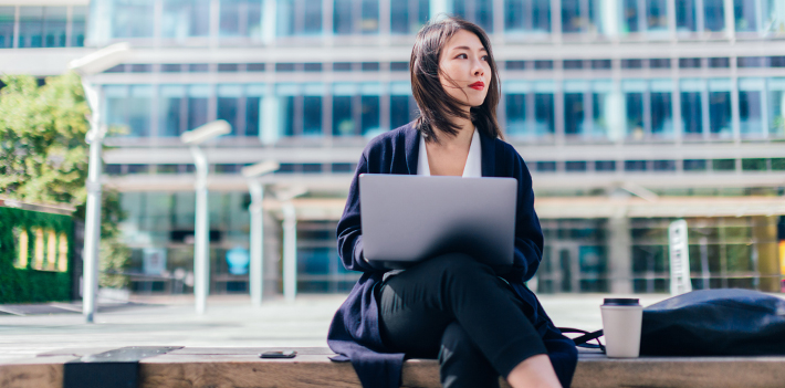 A woman on a laptop outside a high-rise building.