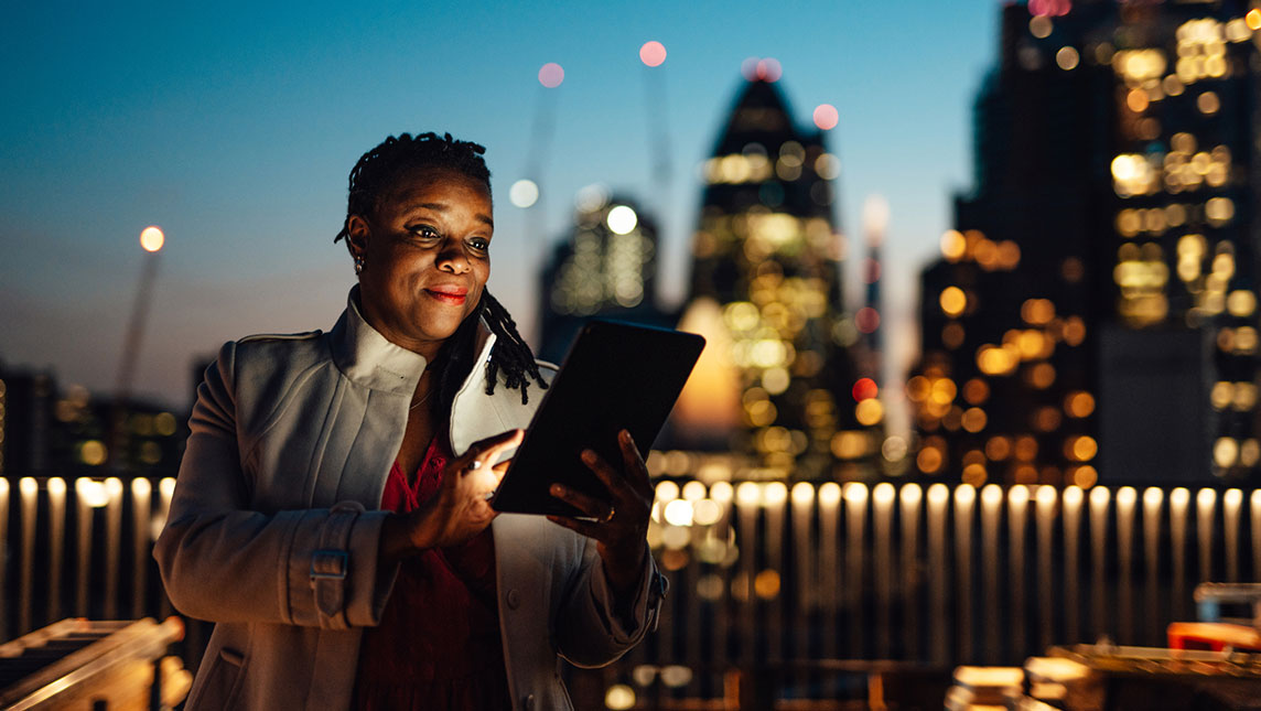 A woman uses a tablet against a city skyline as night draws in.